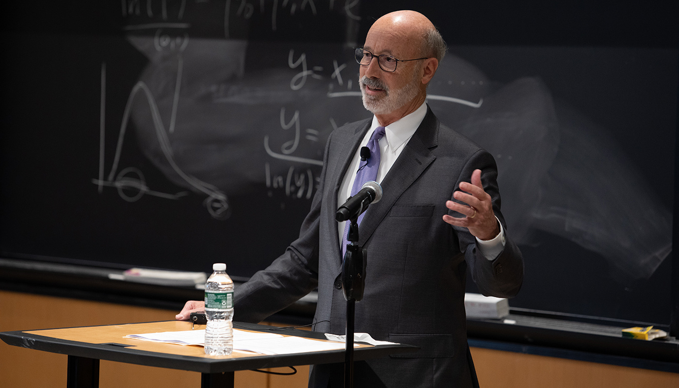 Pennsylvania Governor Tom Wolf speaking in front of MIT chalkboard with equations written on it